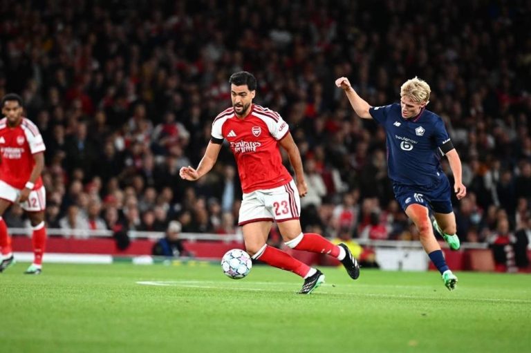 Arsenal player number 23 in red and white dribbles the ball past a Lille defender in a Champions League match at the Emirates.