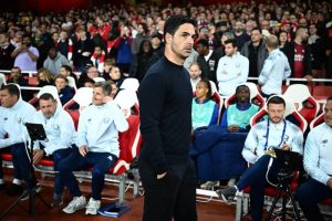 Mikel Arteta in a black sweater standing in a stadium stand, with staff and fans behind him at an Arsenal match.