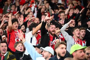 Football fans in red and white scarves cheering loudly and pointing in a crowded stand, matching Arsenal-linked rivalry atmosphere at a Premier League match