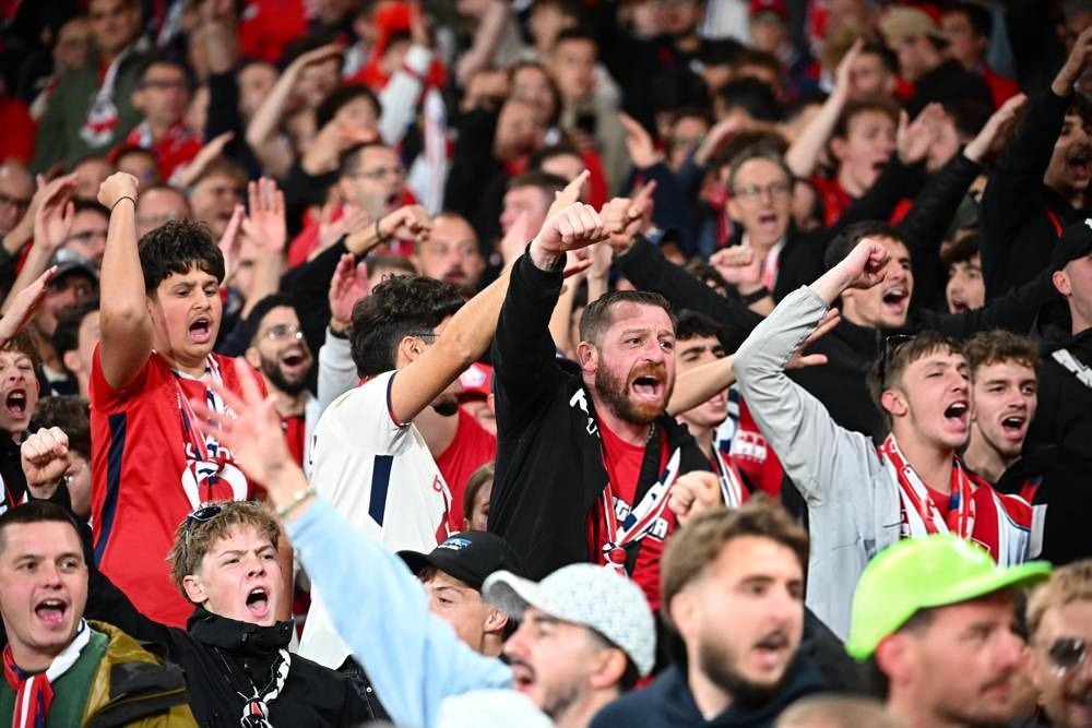 Football fans in red and white scarves cheering loudly and pointing in a crowded stand, matching Arsenal-linked rivalry atmosphere at a Premier League match