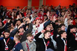 Football supporters in red and white scarves cheer with raised arms and open mouths in a packed stand, likely Arsenal fans at a match.