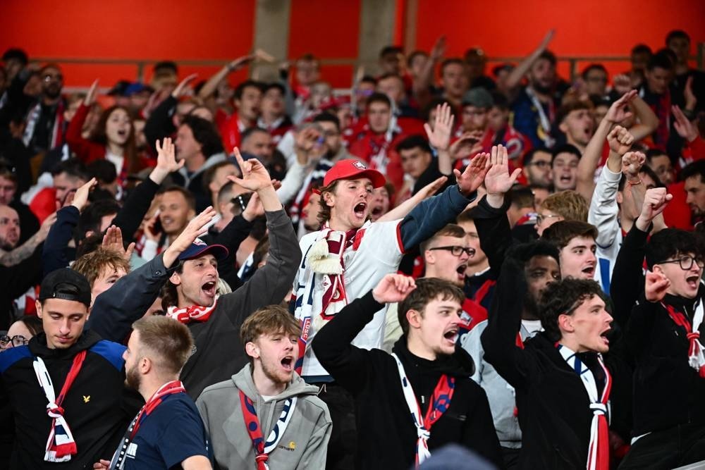 Football supporters in red and white scarves cheer with raised arms and open mouths in a packed stand, likely Arsenal fans at a match.
