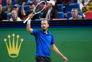 Daniil Medvedev raising his Dunlop racket to fans during a tennis match, wearing a blue Lacoste polo in a stadium.