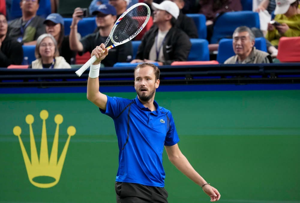 Daniil Medvedev raising his Dunlop racket to fans during a tennis match, wearing a blue Lacoste polo in a stadium.
