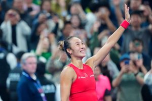 Zheng Qinwen raises her arm and smiles in a red Li-Ning top, with fans cheering and filming behind her on court