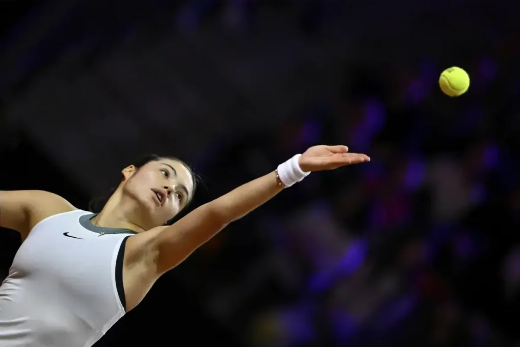 Tennis player in a white Nike sleeveless top and white wristband serving a yellow ball under purple stadium lights