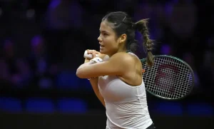 Tennis player in white tank top and braided ponytail holding a racket during a match at an indoor arena, mid-swing with focused expression