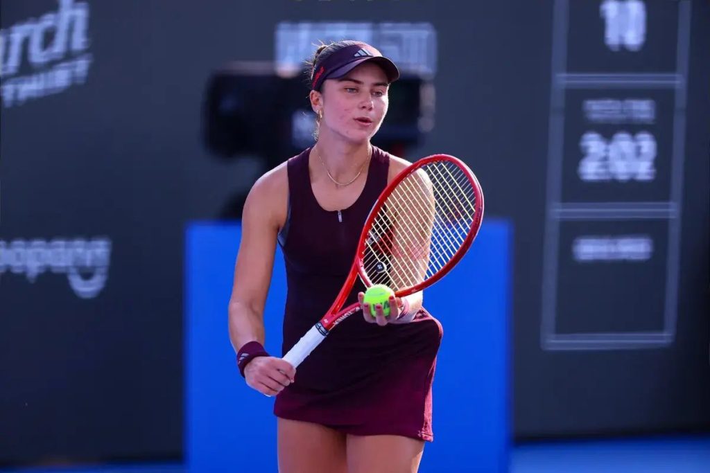 Tennis player in a maroon dress, visor and red racquet holding a yellow ball on a blue court at the Guadalajara tournament