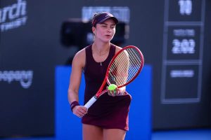 Tennis player in a maroon dress, visor and red racquet holding a yellow ball on a blue court at the Guadalajara tournament