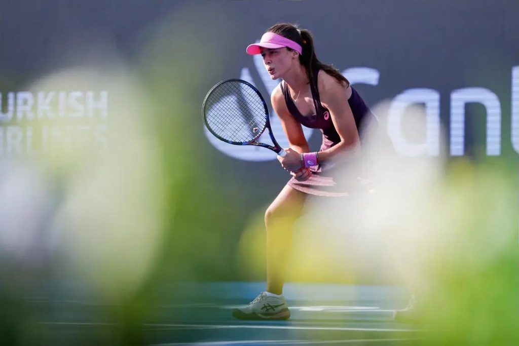 A tennis player in a pink visor and purple outfit, racket ready, crouched on a court with a blurred foreground; sponsor signage behind her.