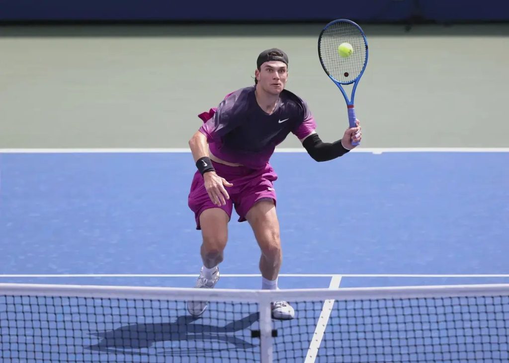 Tennis player in purple Nike outfit and black headband reaches for a forehand volley at the net on a blue court