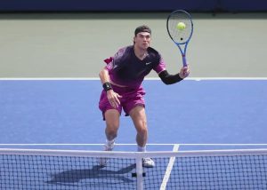 Tennis player in purple Nike outfit and black headband reaches for a forehand volley at the net on a blue court