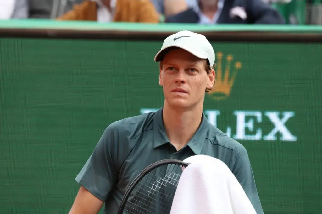 A tennis player in a grey cap and green shirt holding a racket and white towel courtside at a Rolex-sponsored clay court event