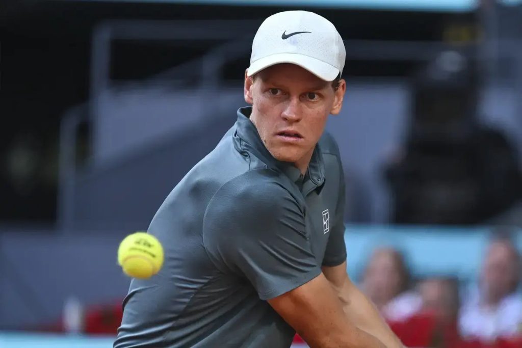 Tennis player in a white Nike cap and grey polo swings a forehand, yellow ball in motion, on a clay court at a match.