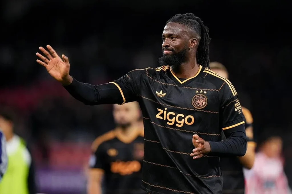 Jogador do Ajax com camisola preta e dourada, de dreadlocks e barba, acena ao público após um jogo, com a equipa de fundo.
