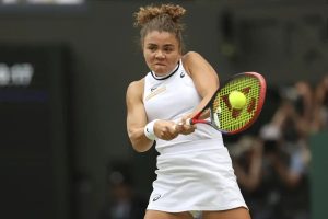 Tennis player in white Asics dress hitting a forehand with a yellow-and-red racket, tennis ball in view, blurred spectators behind on a grass court.