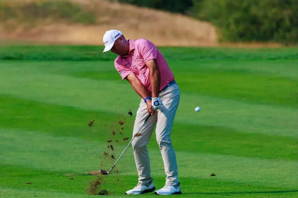 A golfer in a pink polo and white cap hits an iron shot from the fairway, sending turf flying, with the ball in the air.