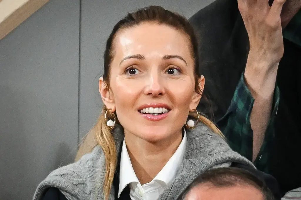 Woman with pulled-back hair, hoop earrings and grey cable-knit vest smiles in a stadium stand, with a man's hand raised behind her.