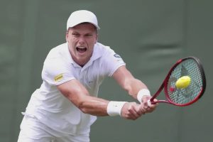 Tennis player in white cap and kit, mid-shout, swinging a red Dunlop racket to return a yellow ball on a green court