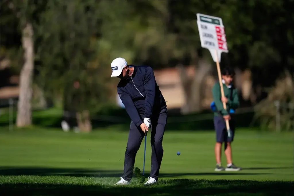 Golfer in navy jacket and white cap with a white glove hits a chip shot on a green fairway, a caddie with a flag behind him