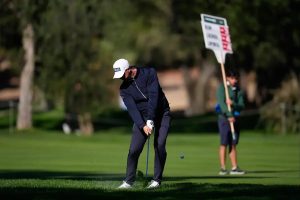 Golfer in navy jacket and white cap with a white glove hits a chip shot on a green fairway, a caddie with a flag behind him