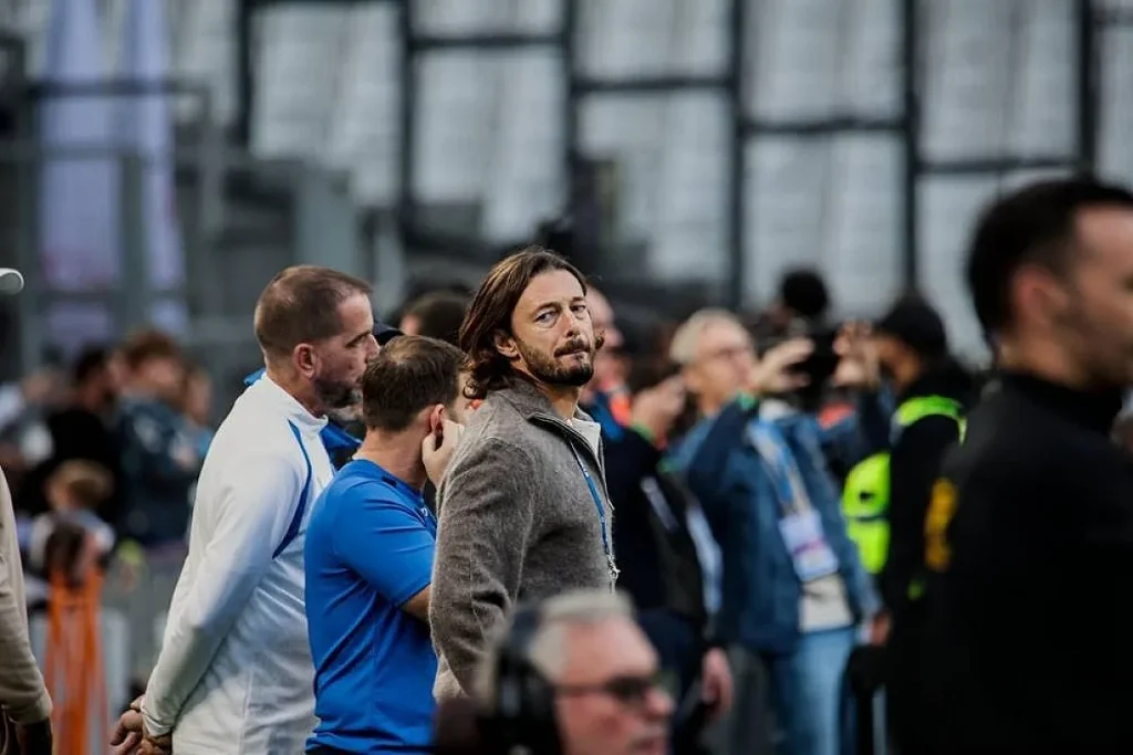 Homem de cabelo comprido e barba, casaco cinzento, num estádio com adeptos e seguranças ao fundo, junto a outros membros da equipa técnica.