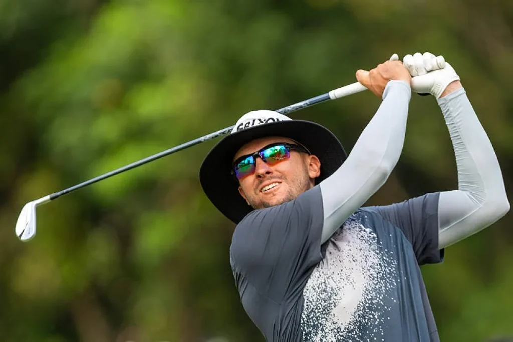 Golfista de chapéu preto e óculos espelhados a fazer um swing com o ferro, em campo verde desfocado.