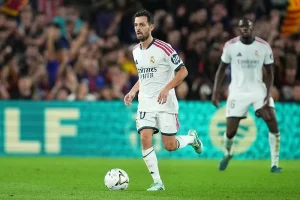 Jogador de branco do Real Madrid, com o número 10, a conduzir a bola num relvado durante um jogo, com adeptos ao fundo.