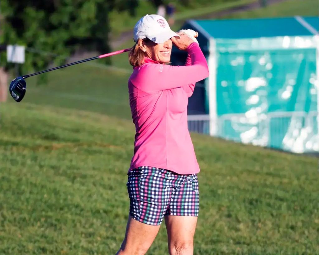 A woman in a pink long-sleeve top, plaid shorts and white cap swings a driver on a grassy golf course, with a teal tent behind her.