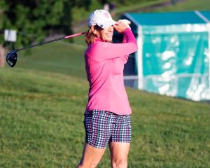 A woman in a pink long-sleeve top, plaid shorts and white cap swings a driver on a grassy golf course, with a teal tent behind her.