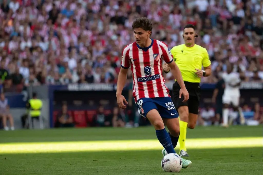 Julian Álvarez in Atlético Madrid red and white stripes dribbling the ball during a match, referee in yellow behind him