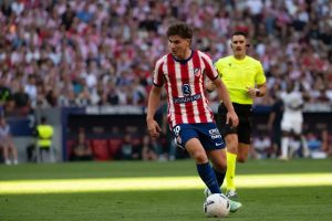 Julian Álvarez in Atlético Madrid red and white stripes dribbling the ball during a match, referee in yellow behind him