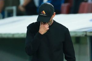 A man in a black cap and black sweatshirt covers his face with his hand on a football touchline, with a blurred stadium behind him.