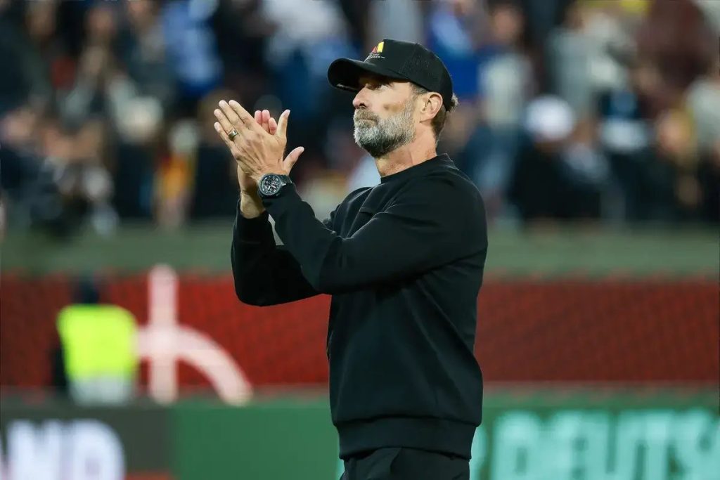 Man in black cap and sweatshirt applauding with hands together, bearded, at a stadium with blurred crowd behind him