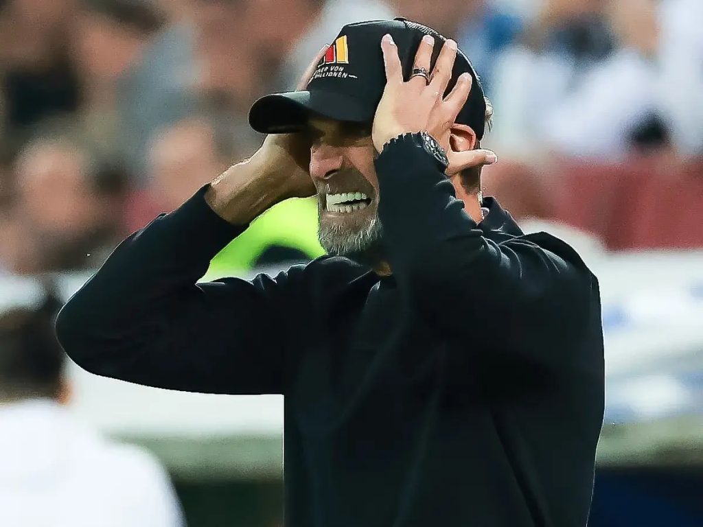 Man in black cap and sweatshirt, hands on head and grimacing in dismay at a stadium, crowd blurred behind him, matching the Germany Nations League defeat story