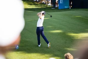 A golfer in a white polo and navy trousers swings a driver on a sunlit fairway, with BMW Championship signage nearby.