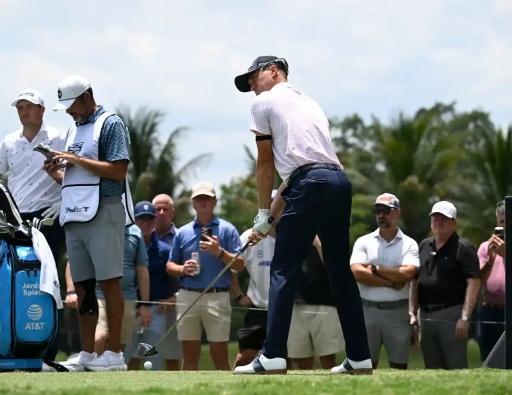 Golfer in pink polo and navy trousers swinging a club on a tee box, caddie with a FedEx bib and crowd watching at a tournament