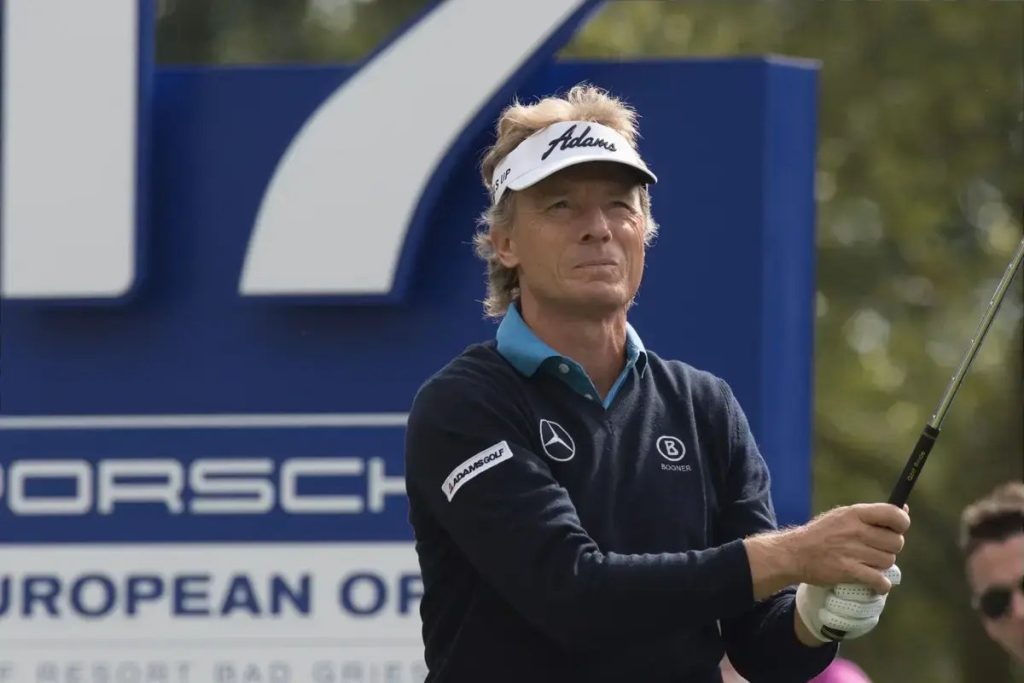 Golfer in a white Adams visor and navy Bogner sweater mid-swing beside a Porsche European Open sign, Bad Griesbach resort
