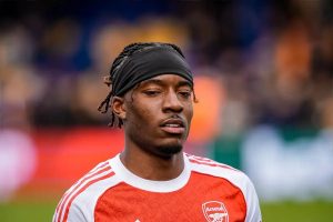 Arsenal player in red and white kit with black headband, looking to the side at a blurred stadium during warm-up