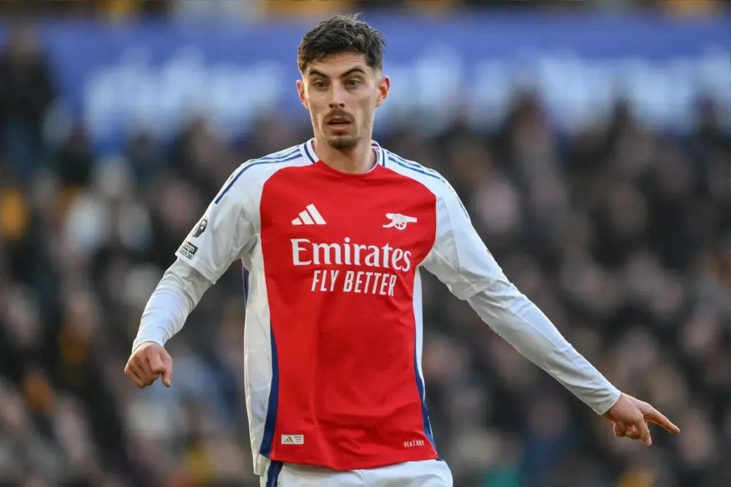 Football player in red and white Arsenal kit with Emirates sponsor, gesturing on the pitch during a Premier League match in a blurred stadium.