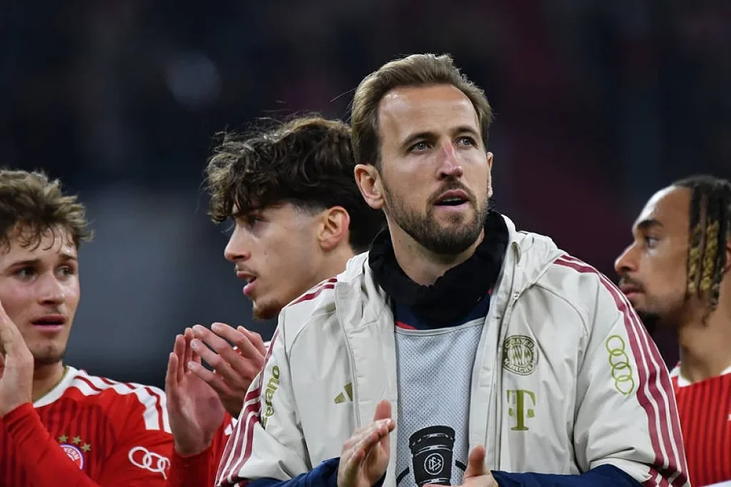 Homem de barba e casaco claro com emblema do Bayern de Munique, aplaudindo ao lado de colegas de equipa de vermelho no estádio.