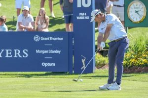 Golfer in white cap and polo swings iron on tee 16, par 4, 462 yards, with spectators and sponsor boards behind