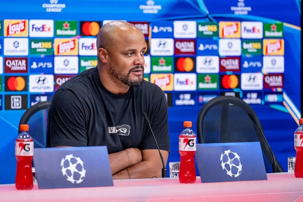 Homem de t-shirt preta sentado numa conferência de imprensa da Liga dos Campeões, de braços cruzados, com microfone e garrafas de bebida à frente.