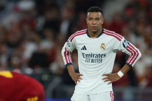 Kylian Mbappe in white Real Madrid kit with hands on hips during a Champions League match, with blurred red-shirted opponent and crowd behind