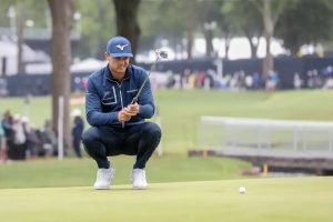 A golfer in a navy quarter-zip and blue Mizuno cap crouches on the green, lining up a putt with his putter as a white ball sits nearby.