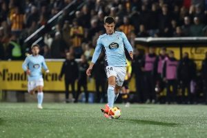 Player in light blue Celta Vigo kit, number 24, dribbling the ball on a wet pitch in a stadium with fans behind.
