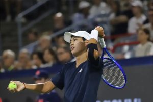 Tennis player in navy Adidas shirt and white cap, holding a yellow ball and blue racket, preparing to serve at a night match.