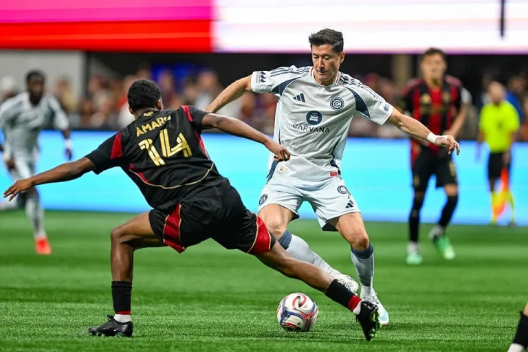 Jogador de branco com equipa do Chicago Fire disputa a bola com defesa de preto e vermelho nº 14, Amari'i, em estádio.