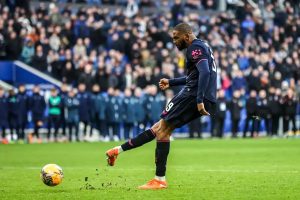 Footballer in navy kit with number 9 striking a penalty at a stadium, crowd and players behind. Article subject is Brian Brobbey.