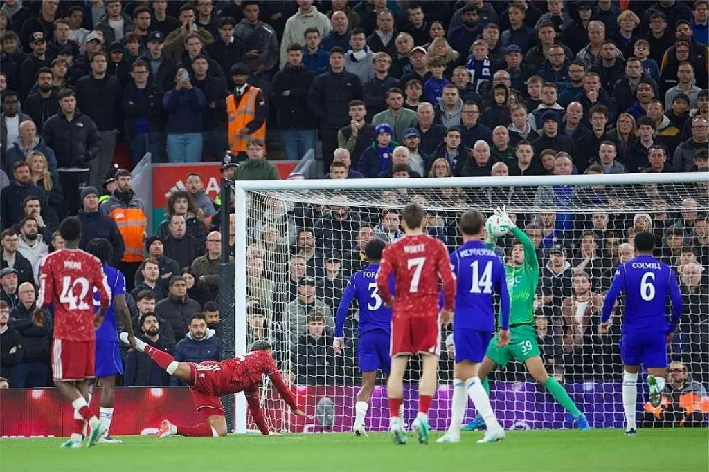 Guarda-redes do Chelsea (camisola verde, n.º 30) defende um penálti de jogador do Liverpool (equipamento vermelho, n.º 7) num estádio lotado